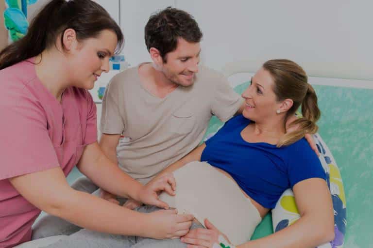 Femme souriante accompagné de son mari, allongée sur un lit et se faisant soigner par une infirmière