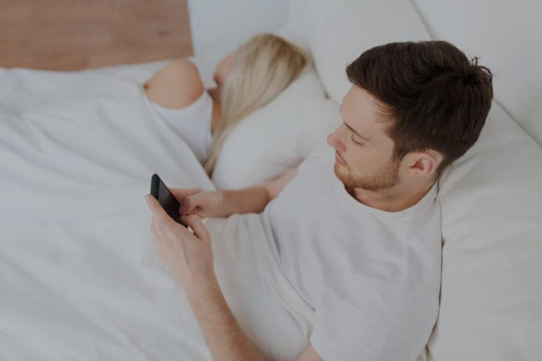 Homme brun allongé dans sont lit qui regarde son téléphone portable à côté d'une femme blonde qui dort