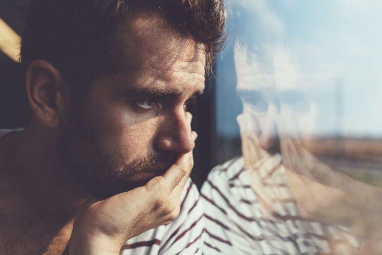 homme brun qui a la main sur la bouche et qui regarde l'horizon par la fenêtre